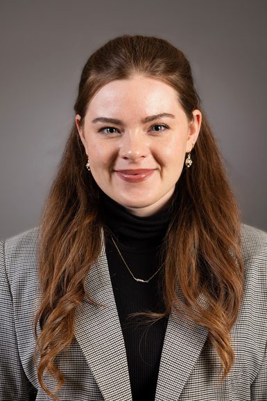 A woman looking straight to camera with long brown hair wearing a black jumper with a grey patterned blazer.