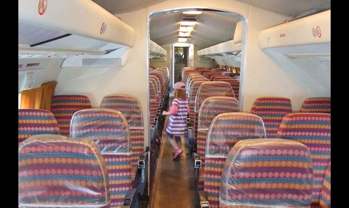 The interior of an airplane with two seats on each side of the aisle. The seats are covered in a striped red, pink, blue, and yellow upholstery and there is a child walking down the aisle.