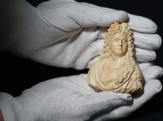 A pair of hands wearing white gloves holding a small ivory statue of a man from the shoulders up with long curly hair and a draped shirt