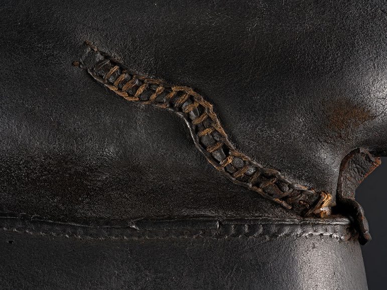 Detail of long black 17th century Jack boots showing a stitched repair against a black background.