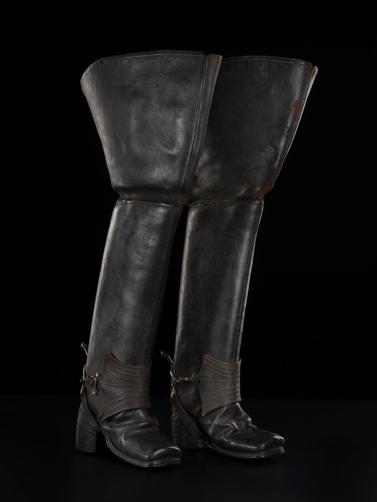 A pair of long black 17th century Jack boots propped up against a black background.