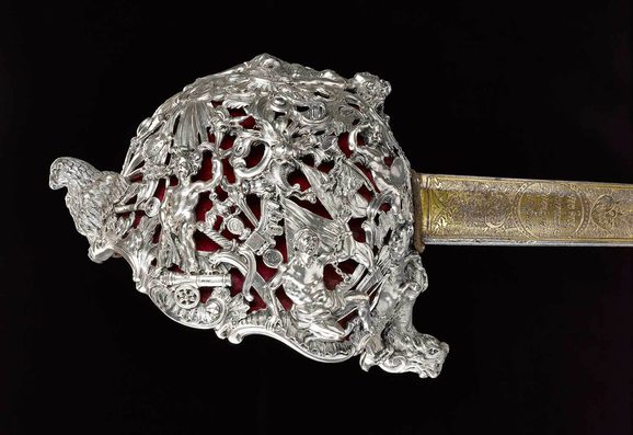 Silver sword hilt densely packed with designs of cannons, flags, swords, eagles, swans and swirls. A gold blade detail is visible to the right hand side of the image. Set against the black background.