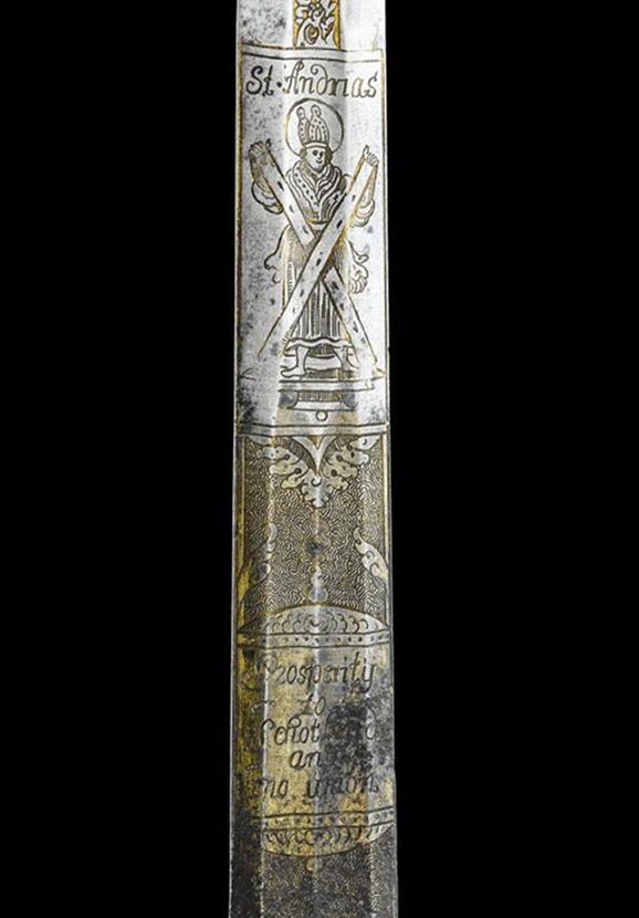 A close up detail of a sword, depicting a man dressed in a robe with a long hat, holding two planks creating a shape of an X. 'St Andrias' inscribed above the head of the man.