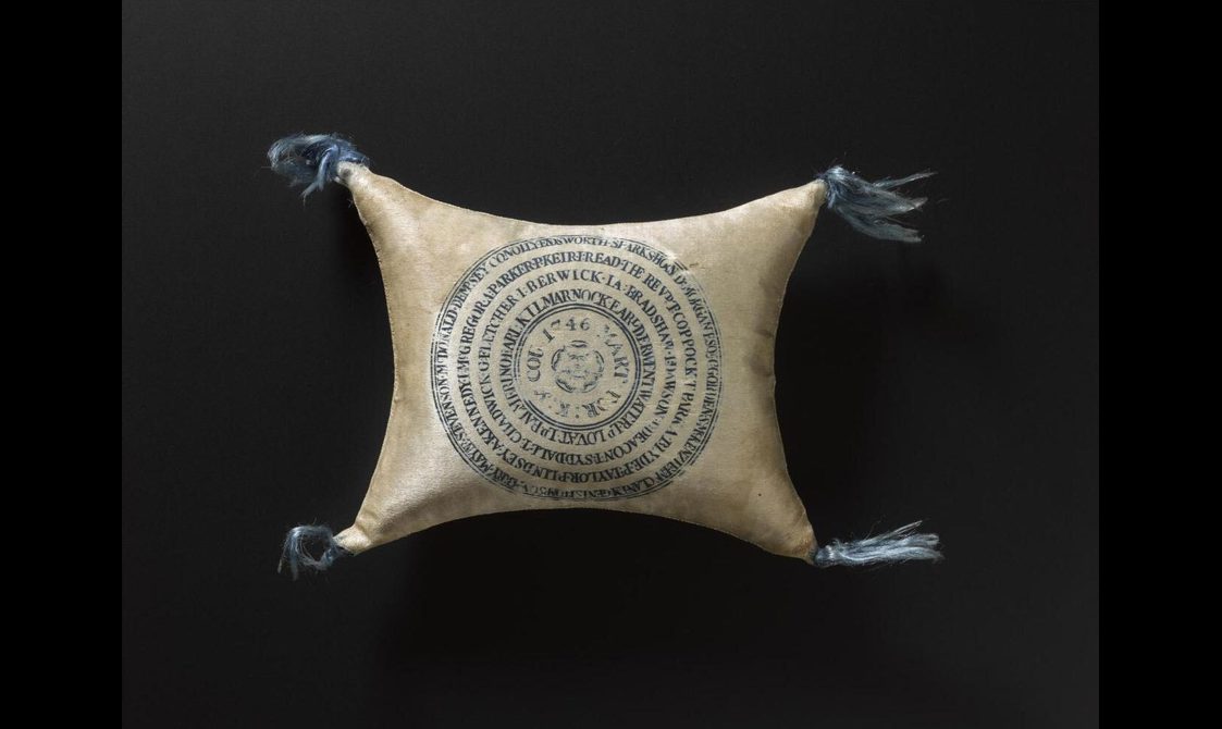 A cream colour velvet pin cushion with slightly faded blue tassels attached to each corner. Multiple names are printed in the center of the cushion in a circular manner. Set against a black background.