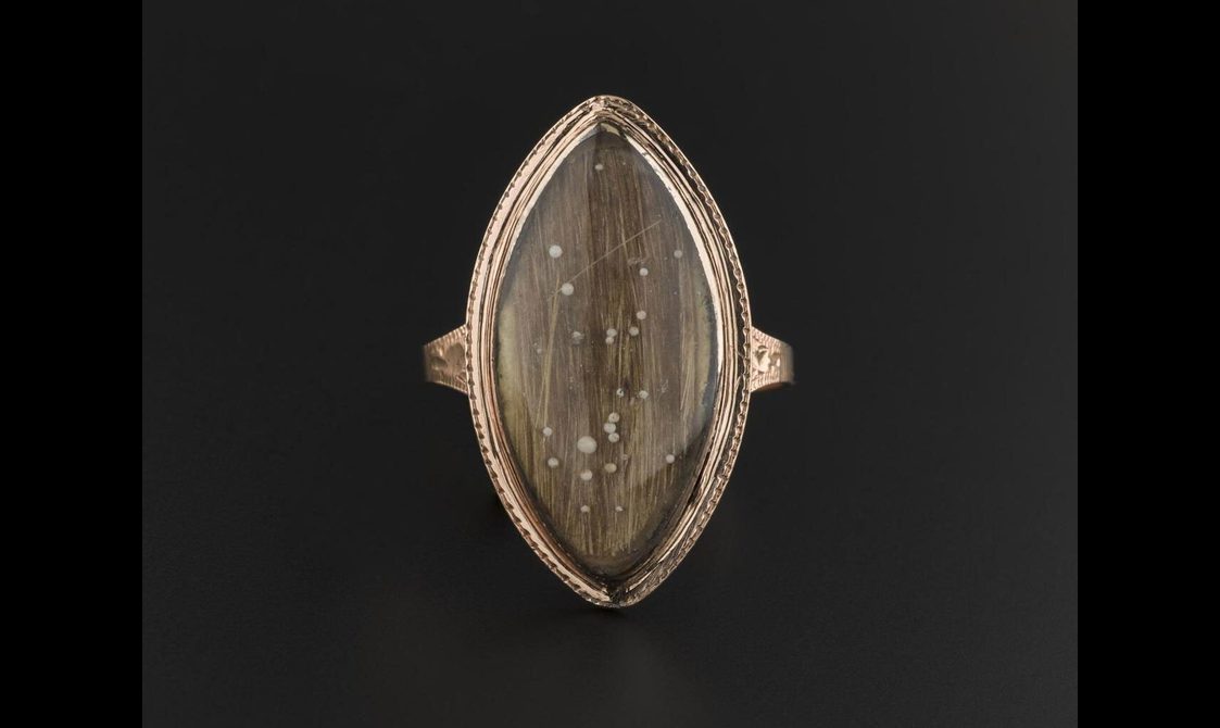 A gold oval shaped ring with sharpened corners set against a black background. A lock of light brown hair is set inside, with a few white speckles.
