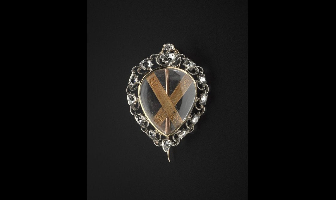 An upside down drop-shaped glass brooch with two strands of light brown hair shaped in a shape of an 'X' set against the black background. Silver and diamond decoration set around the glass container.