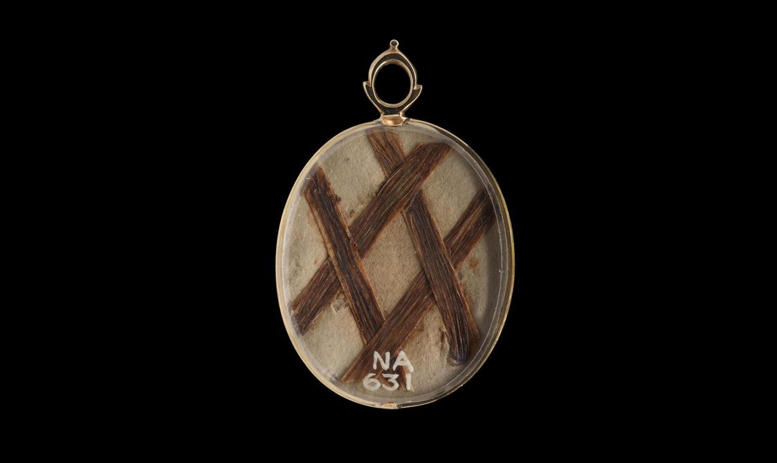 A gold oval shaped locket with four strands of brown hair set inside in a shape of a diagonal lattice plait. Set against the black background.