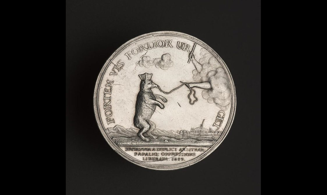 A silver medal with inscriptions to the edges, a bear stood on its rear legs with a lead attached to the nose. Two hands attached to a cloud, one holding a lead and the other a baton.