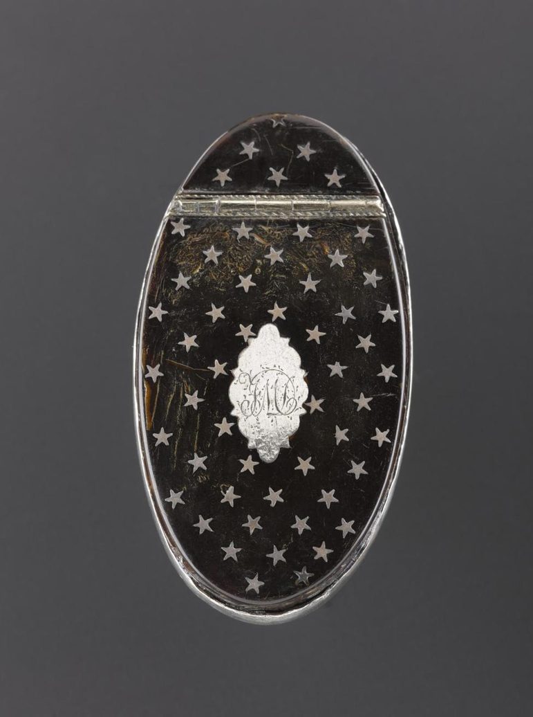 An oval black tortoiseshell snuff box lid pictured from above. The box is decorated with five pointed stars. In the center, a silver oval engraved with initials 'FM'.