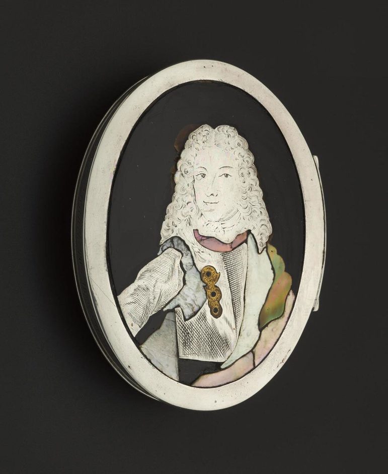 A black oval shaped tortoiseshell oval box with white ridge set against the black background. A white portrait of a woman wearing a curly shoulder length wig, dressed in a white jacket with golden brooch.
