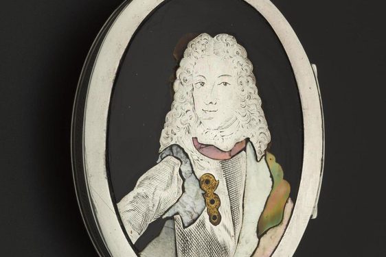 A black oval shaped tortoiseshell oval box with white ridge set against the black background. A white portrait of a woman wearing a curly shoulder length wig, dressed in a white jacket with golden brooch.