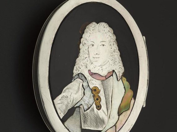 A black oval shaped tortoiseshell oval box with white ridge set against the black background. A white portrait of a woman wearing a curly shoulder length wig, dressed in a white jacket with golden brooch.