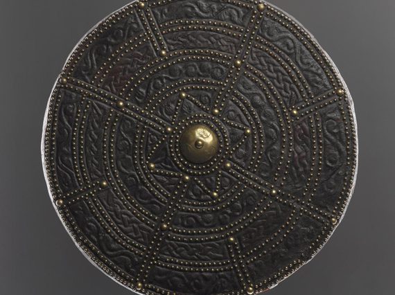 A round leather shield, embossed with decorated with pattern and small brass pegs throughout. The middle of the shield is decorated with a large brass peg.