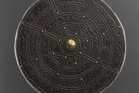 A round leather shield, embossed with decorated with pattern and small brass pegs throughout. The middle of the shield is decorated with a large brass peg.