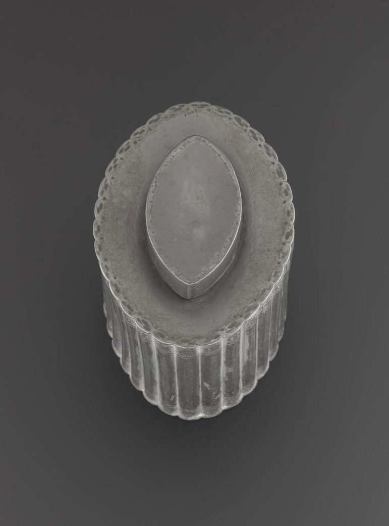 A grey oval shaped tea caddy with a smaller oval shaped lid atop. Set agaisnt grey background.