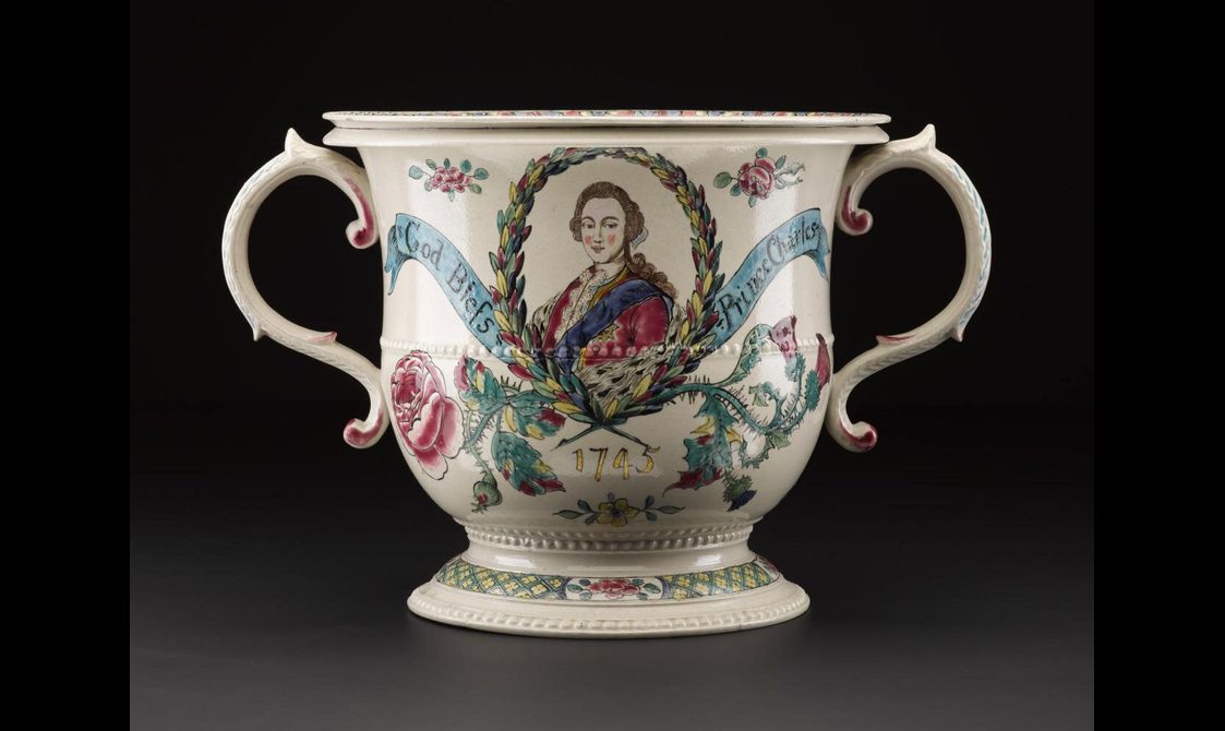 A white porcelain mug with two handles to the sides. An oval portrait of a man wearing a red coat, blue ribbon over the left shoulder, and a brown wig, situated in the center of the mug. Floral motifs surround the portrait and a blue ribbon going across with inscription "God Bless Prince Charles""