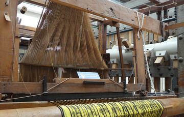 A large wooden loom with a lot of strands bunched together with a yellow and black cloth being woven.