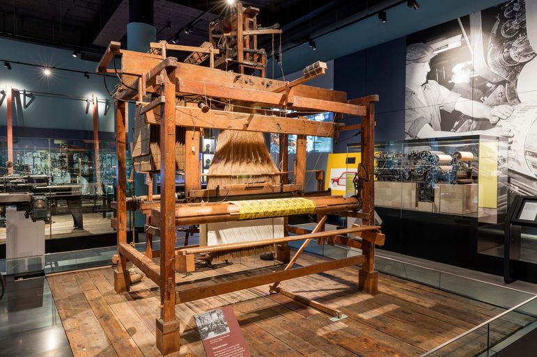 A large wooden loom sits on a wooden plinth in a museum setting