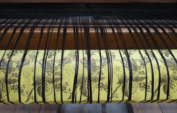 A close up of a wooden loom showing thin black strands coming over a yellow and black patterned cloth