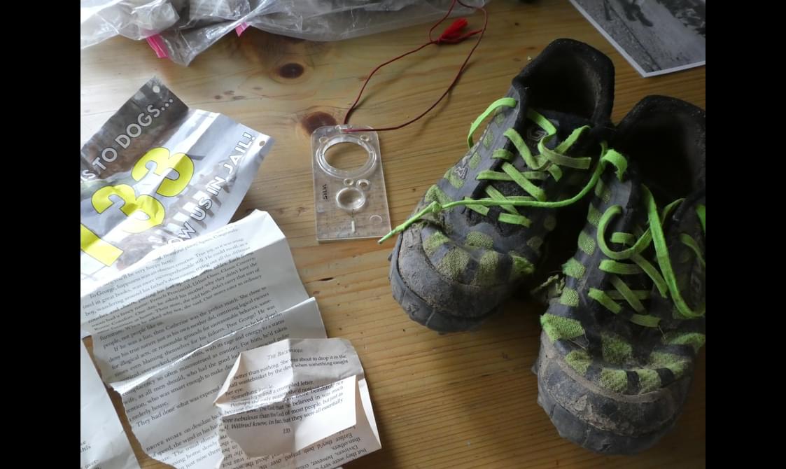 A pair of muddy green and black running shoes, compass, race number, plastic bags, and torn book page from the 2024 Barkley Marathon.