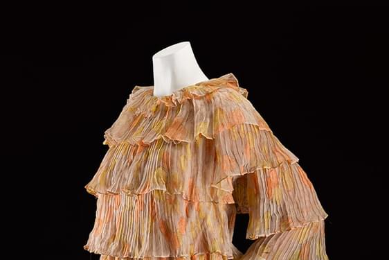 A mannequin wearing a beige and orange patterned mini dress with tiered silk chiffon layers and three quarter length sleeves.