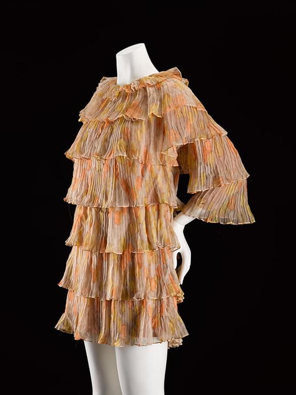 A mannequin wearing a beige and orange patterned mini dress with tiered silk chiffon layers and three quarter length sleeves.