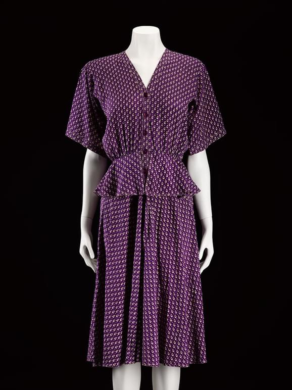 A mannequine wearing a patterned purple dress with short sleeves, buttons down the front and a peplum at the waist.