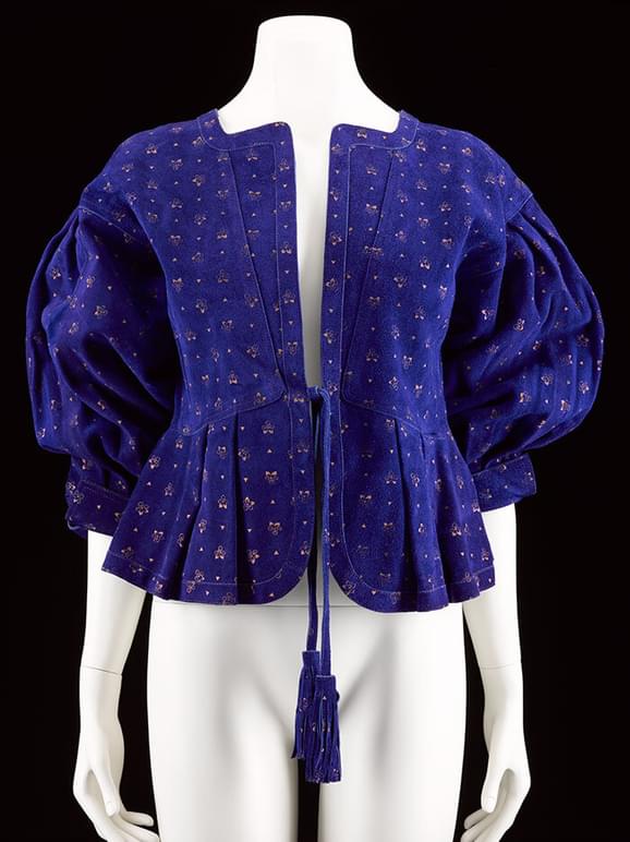 A mannequin wearing a royal blue suede top with a gold printed pattern and voluminous sleeves with a tassled waist tie.