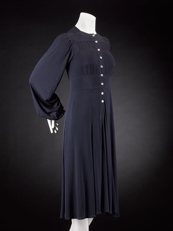 A mannequin wearing a navy knee-length dress with white buttons from the neck to the waist, and long voluminous sleeves.