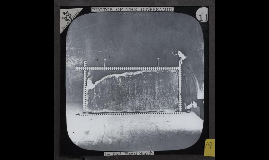 A black and white negative slide of a woman in a long dark dress standing next to a large rock that has measuring sticks around it. The slide has stickers and labels around the outside.