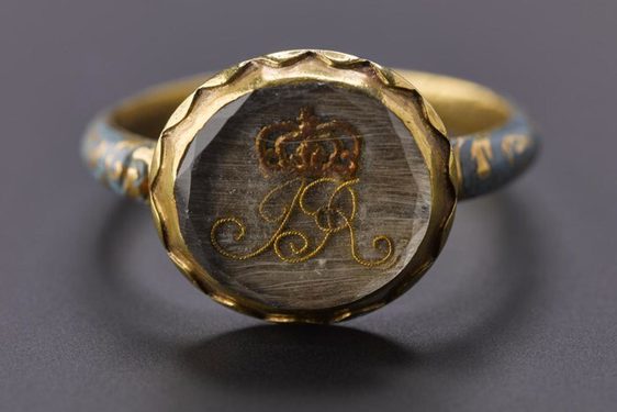 Close up of a circle shaped ring, with a clear gem stone. "JR" initials formed from a light brown thread and a red brown crown design above it.