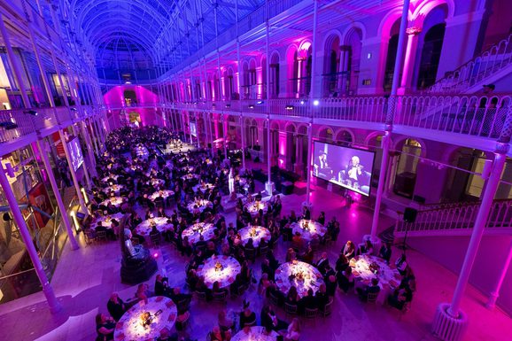 A multi-level gallery space with balconies running around the outside. The gallery is filled with people sitting at round tables listening to two people talk. The gallery is illuminated with blue, purple, and pink lights.