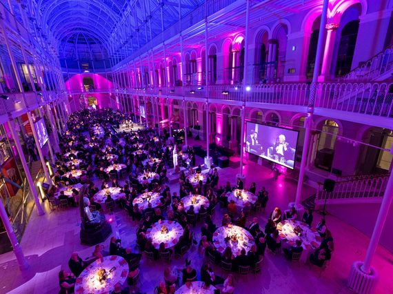 A multi-level gallery space with balconies running around the outside. The gallery is filled with people sitting at round tables listening to two people talk. The gallery is illuminated with blue, purple, and pink lights.