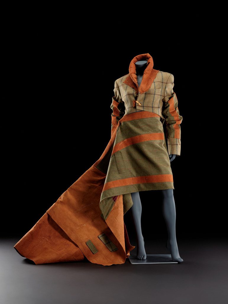 A mannequin wearing a tan jacket with a large orange collar and orange and green sleeves, and a green and orange wide-striped asymmetrical skirt with a train to the side.