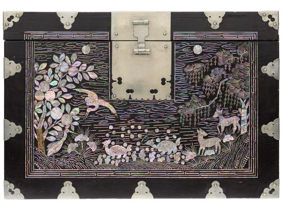 The top of a black bock with pearl embellishments depicting trees, birds, and animals. There is a large latch or hinge in the top middle.