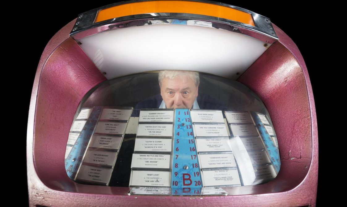 A colour photograph of a man with white hair and a blue-collared shirt, looking through the window of a jukebox from the back. In the front is the card display of records in the jukebox.