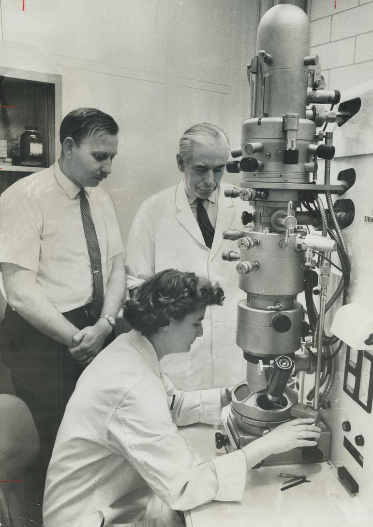 A black and white image of a woman in a lab coat operating a large microscope, with two men standing nearby observing.