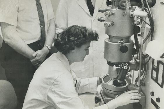 A black and white image of a woman in a lab coat operating a large microscope, with two men standing nearby observing.