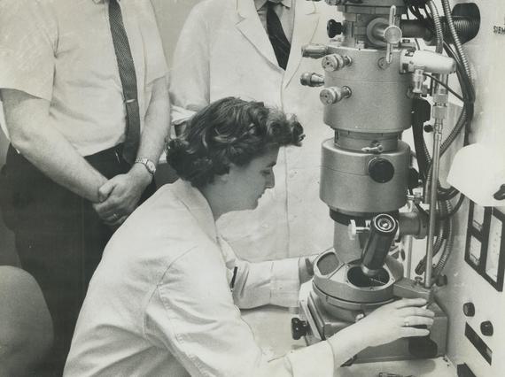 A black and white image of a woman in a lab coat operating a large microscope, with two men standing nearby observing.
