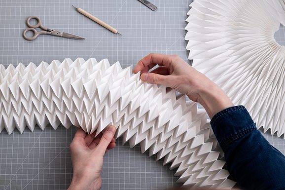 Paper folded intricately on a craft grid board, making a long rectangular structure and a circular structure in the background.