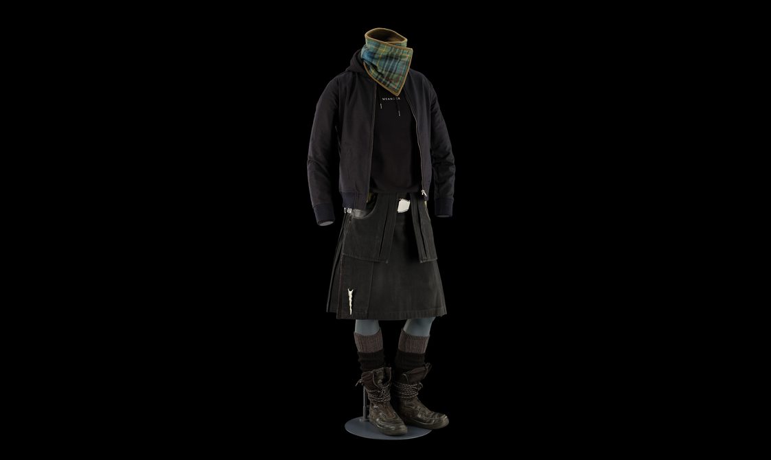 A mannequin wearing a black denim kilt, a black hoodie, a black jacket, and a green and blue tartan neck scarf.