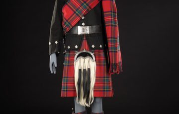A red man's tartan kilt suit with draped red tartan fabric across left shoulder and a black army jacket.
