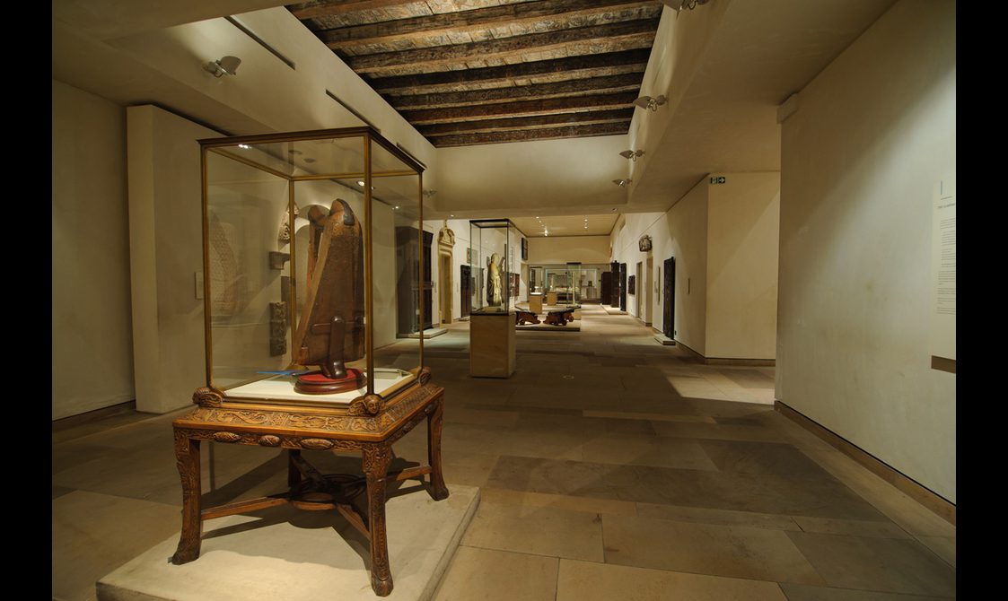 A long, narrow, low-lit gallery space with stone floors and a wooden ceiling. There are free-standing glass display cases along the floor.