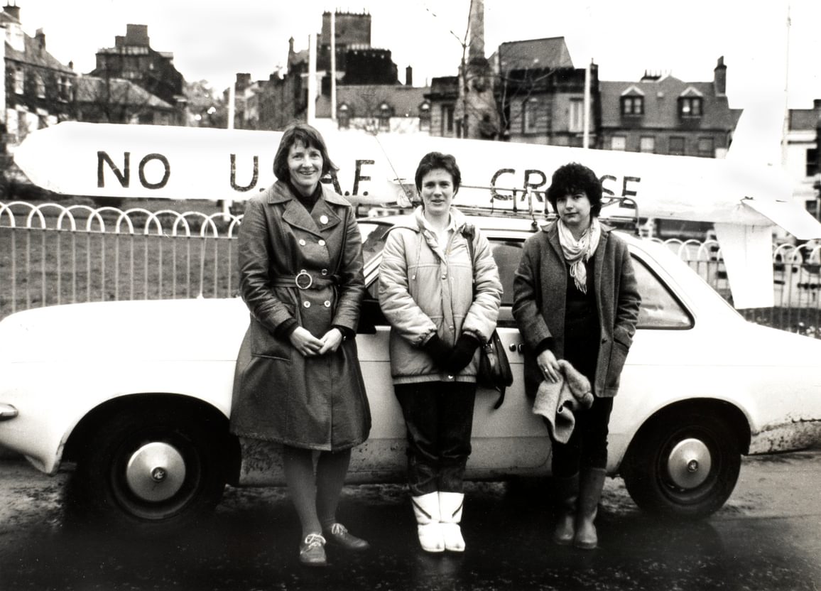 Kristin Barrett and two other women standing in front of a white car, with a cardboard cruise missile with slogan in the background.