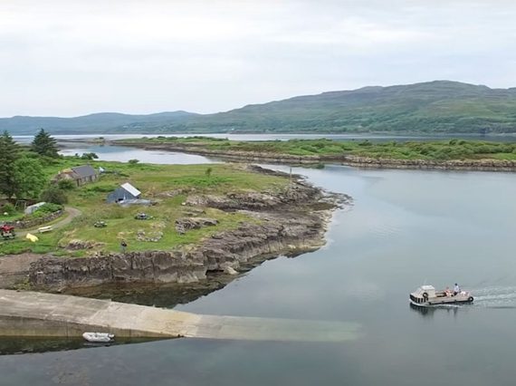 A boat on an inlet of water pulls into a jetty where there is a white house. There are green hills in the background.