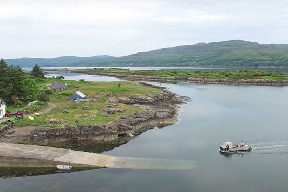 A boat on an inlet of water pulls into a jetty where there is a white house. There are green hills in the background.