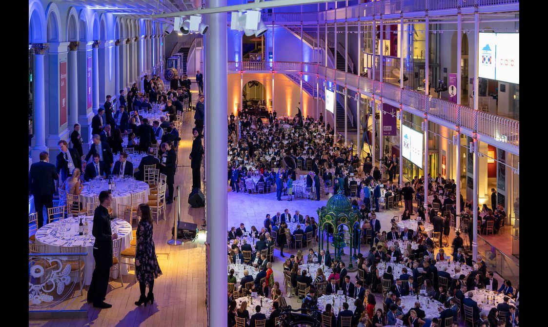 People at a dinner reception in a multi-level gallery space. The space is illuminated with blue and yellow lights.