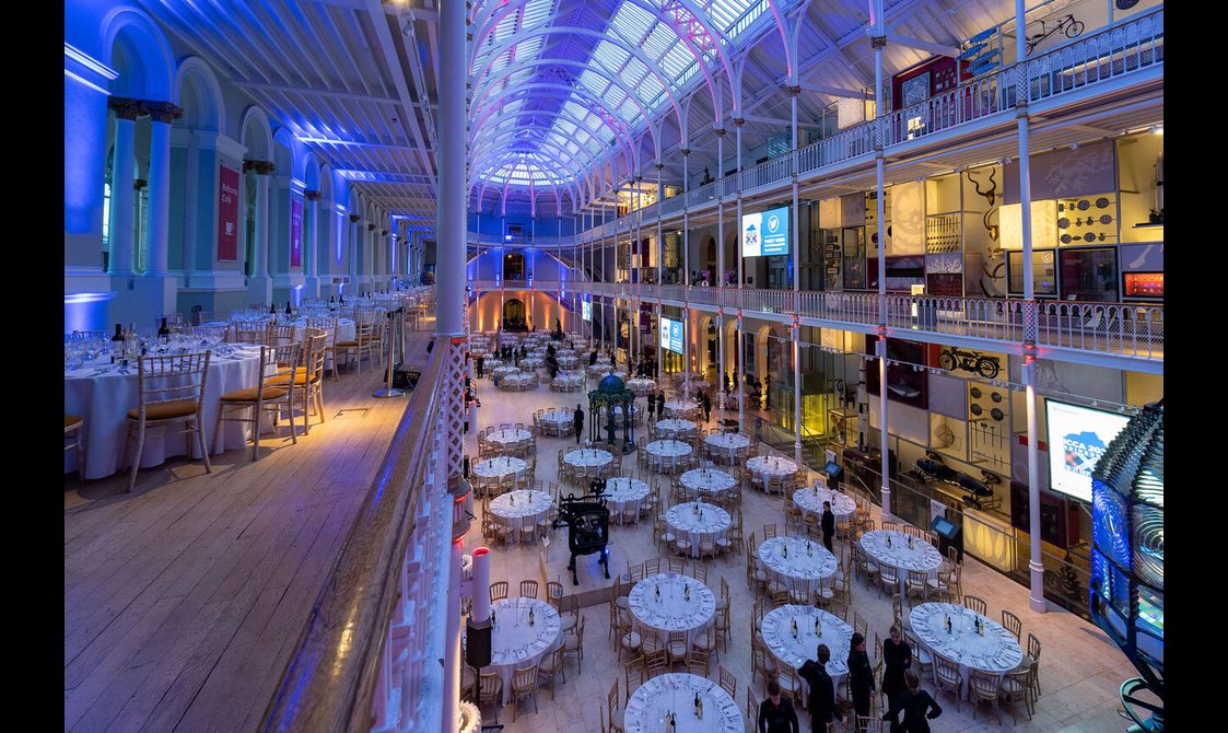 Tables set up for a dinner reception in a multi-level gallery space. The space is illuminated with blue and yellow lights.