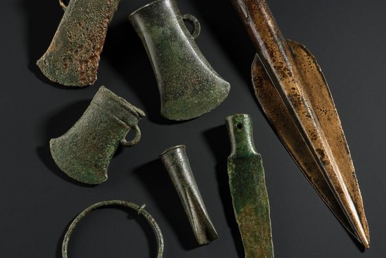 Three axe heads, three spears, and two large rings all made of discoloured bronze sitting on a black background
