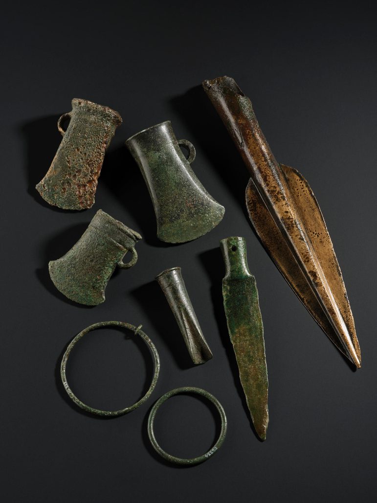 Three axe heads, three spears, and two large rings all made of discoloured bronze sitting on a black background